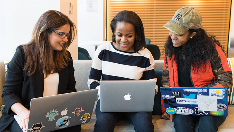 Three women of color sitting together on a couch working from laptops.